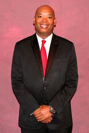 Man in a black suit with a red tie standing against a pink backdrop, hands clasped.