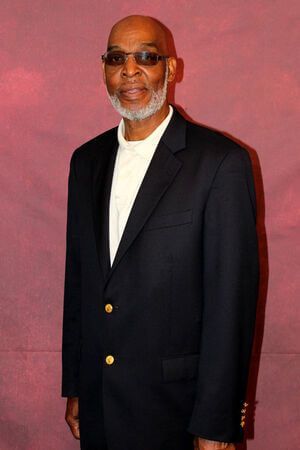 Man in a black blazer and sunglasses standing against a pink wall