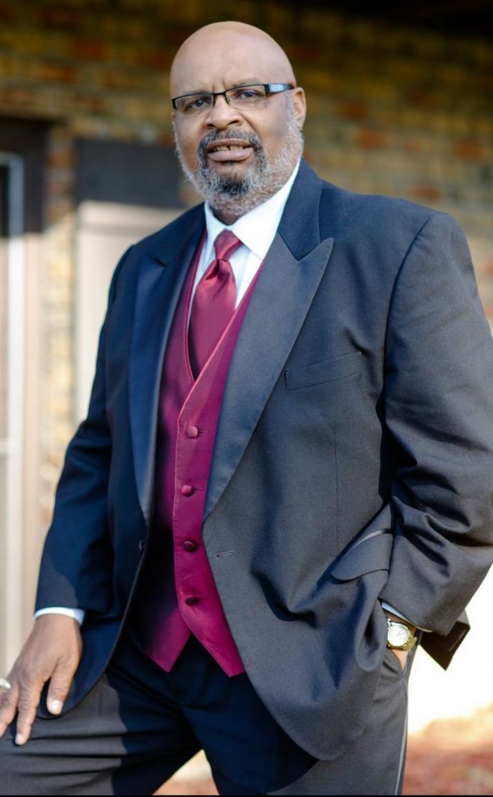 Man in a dark suit with burgundy vest and tie standing outdoors, leaning on a ledge