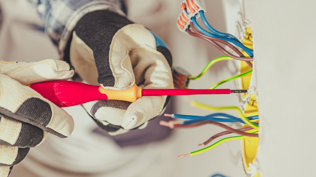 Gloved hands use a red screwdriver to work on colorful electrical wires protruding from a yellow wall junction box.