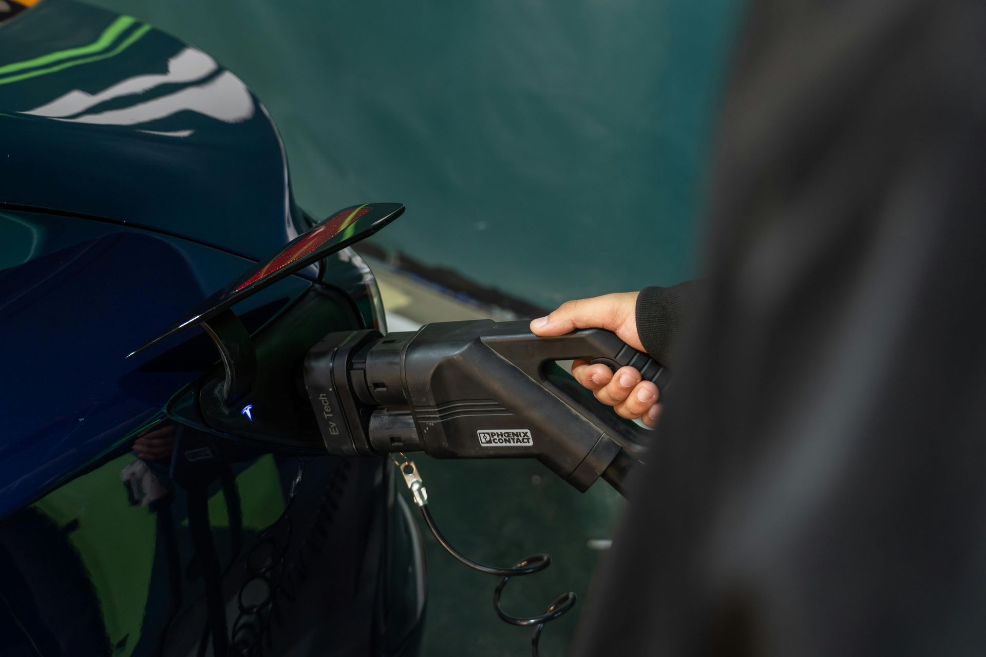 A hand holds a black electric vehicle charging plug connected to the charging port of a teal and dark blue vehicle.