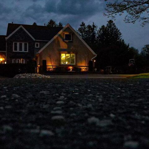 Dusk view of a house with glowing porch lights, seen low from a dark stone driveway.