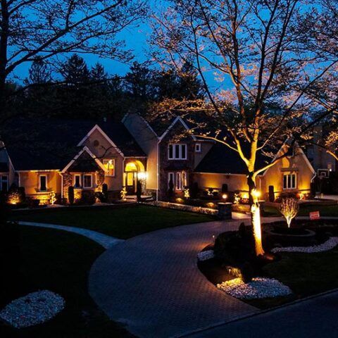 Elegant house lit warmly at dusk, with a curved driveway and illuminated tree in the front yard