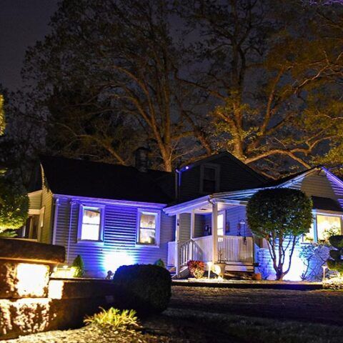 Suburban house at night lit with blue and warm white lights, with trees and a front porch