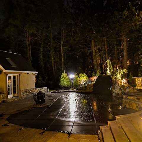 Nighttime backyard patio with wet stone tiles, lit trees, and a house on the left