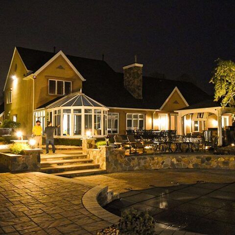 Lit stone house and patio at night, warmly illuminated with a glass gazebo and steps leading up.