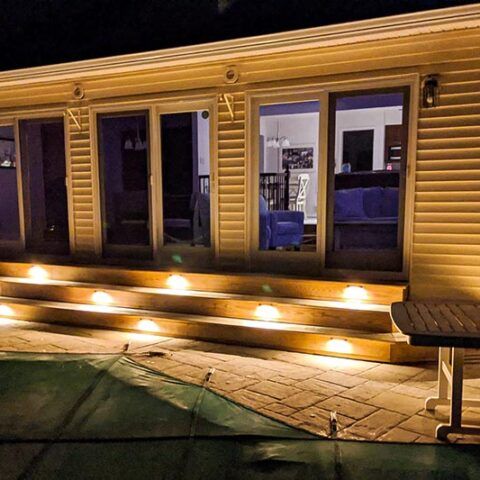 Warmly lit wooden deck at night with steps, glass doors, and a patio table beside a house.