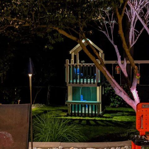 Night garden with a small wooden playhouse lit by blue lights, beside a tree and lawn.