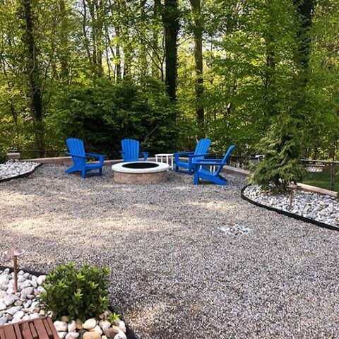 Blue Adirondack chairs around a fire pit on a gravel patio in a wooded backyard.