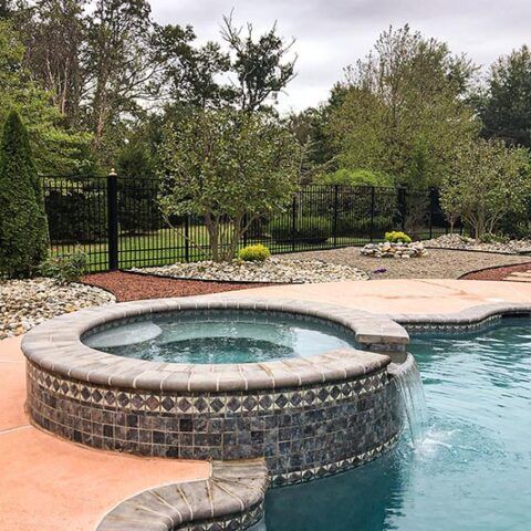 Backyard pool with raised circular hot tub, stone coping, and trees behind a black fence