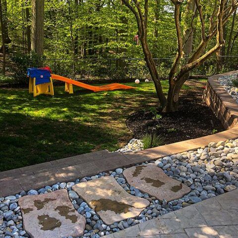 Backyard garden with a small orange slide, trees, stone path, and pebble edging around a patio