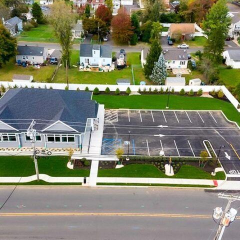 Aerial view of a building with an empty parking lot, street frontage, and houses behind it.