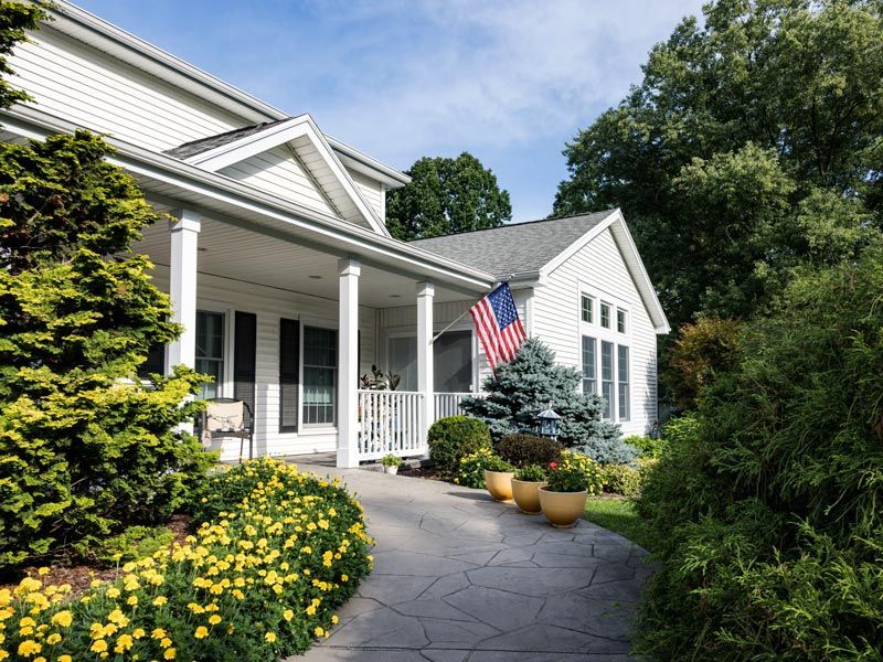 Suburban home with white porch, American flag, and flower-lined walkway on a sunny day
