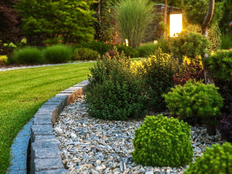 Curved stone garden border with green shrubs, mulch, and a lit lamp in a landscaped yard