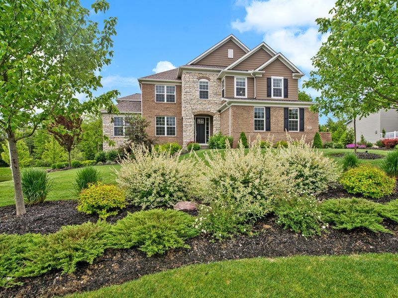 Brick suburban house with manicured shrubs and trees in a landscaped front yard under a blue sky