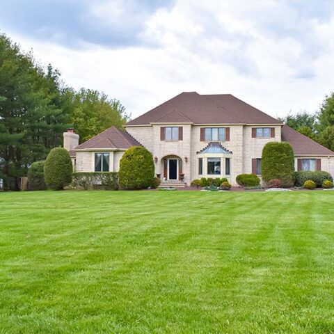Large two-story brick house with a manicured lawn, shrubs, and trees under a cloudy sky