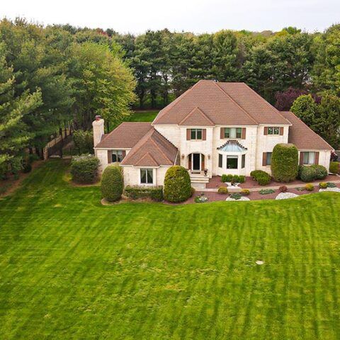 Large beige two-story house with a brown roof, set on a green lawn beside trees