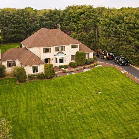 Large beige suburban house with a red roof, driveway, and expansive green lawn beside trees