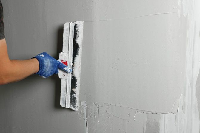 Person wearing blue gloves smoothing gray plaster on a wall with a trowel
