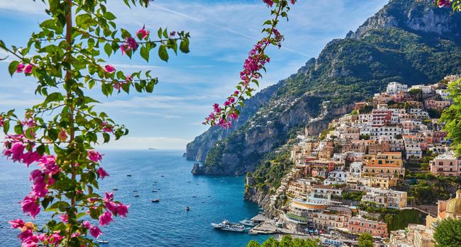 Città costiera sul fianco di una montagna, mare azzurro, fiori rosa, giornata di sole.