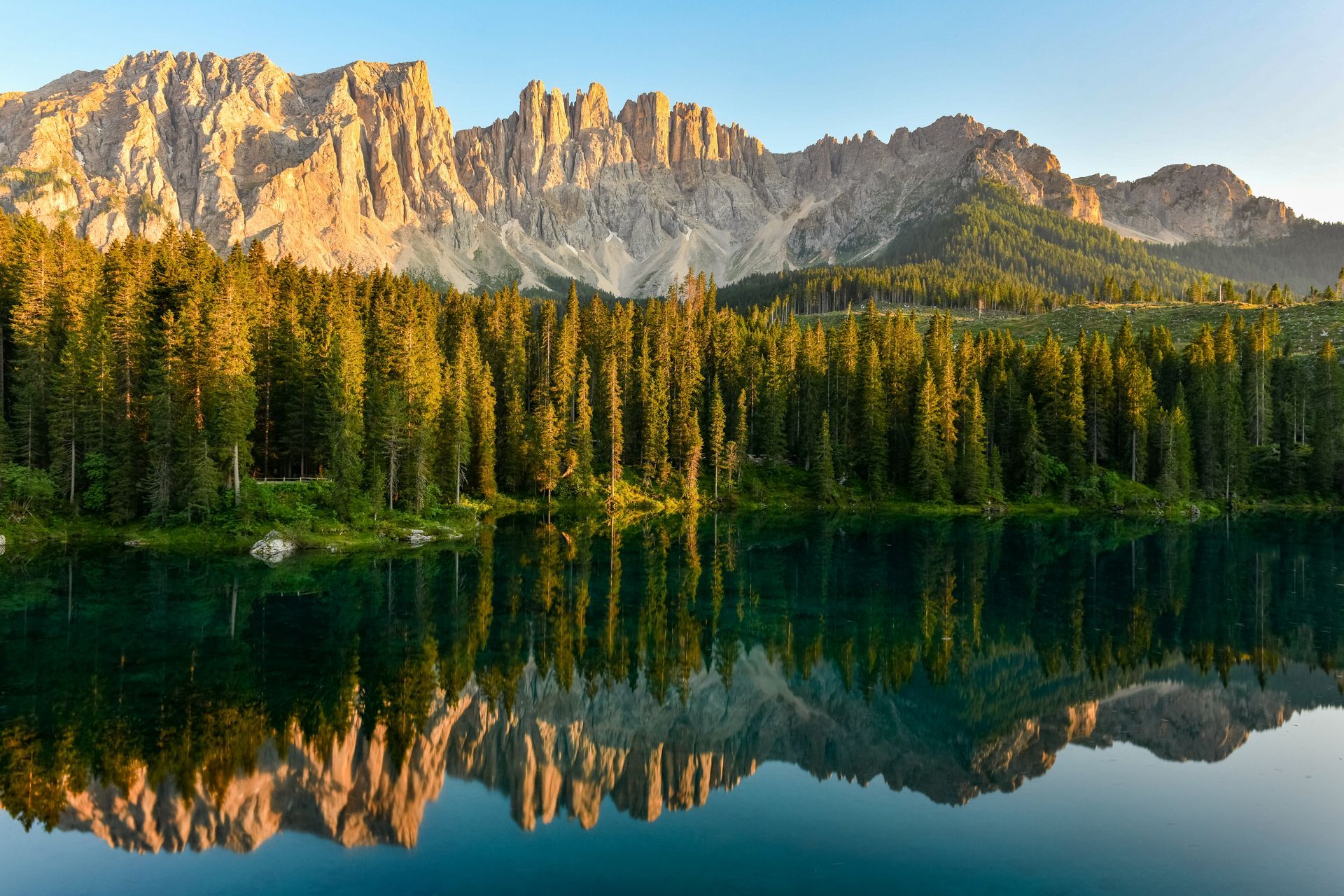 Foresta di alberi sempreverdi riflessa in un lago immobile, con una catena montuosa frastagliata sullo sfondo.