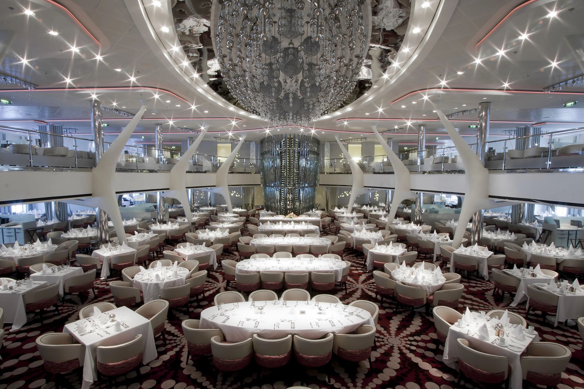 Ampia sala da pranzo su più livelli su una nave da crociera, con numerosi tavoli apparecchiati per il pasto, moquette a motivi geometrici e grande lampadario.