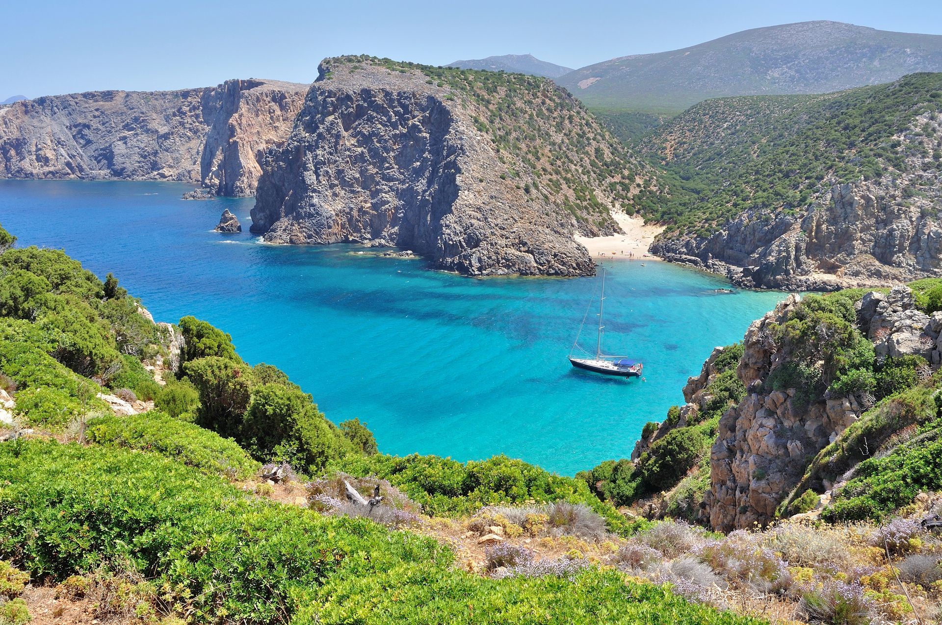 Mare azzurro e scogliere rocciose, con una piccola barca, circondati dal verde della vegetazione.