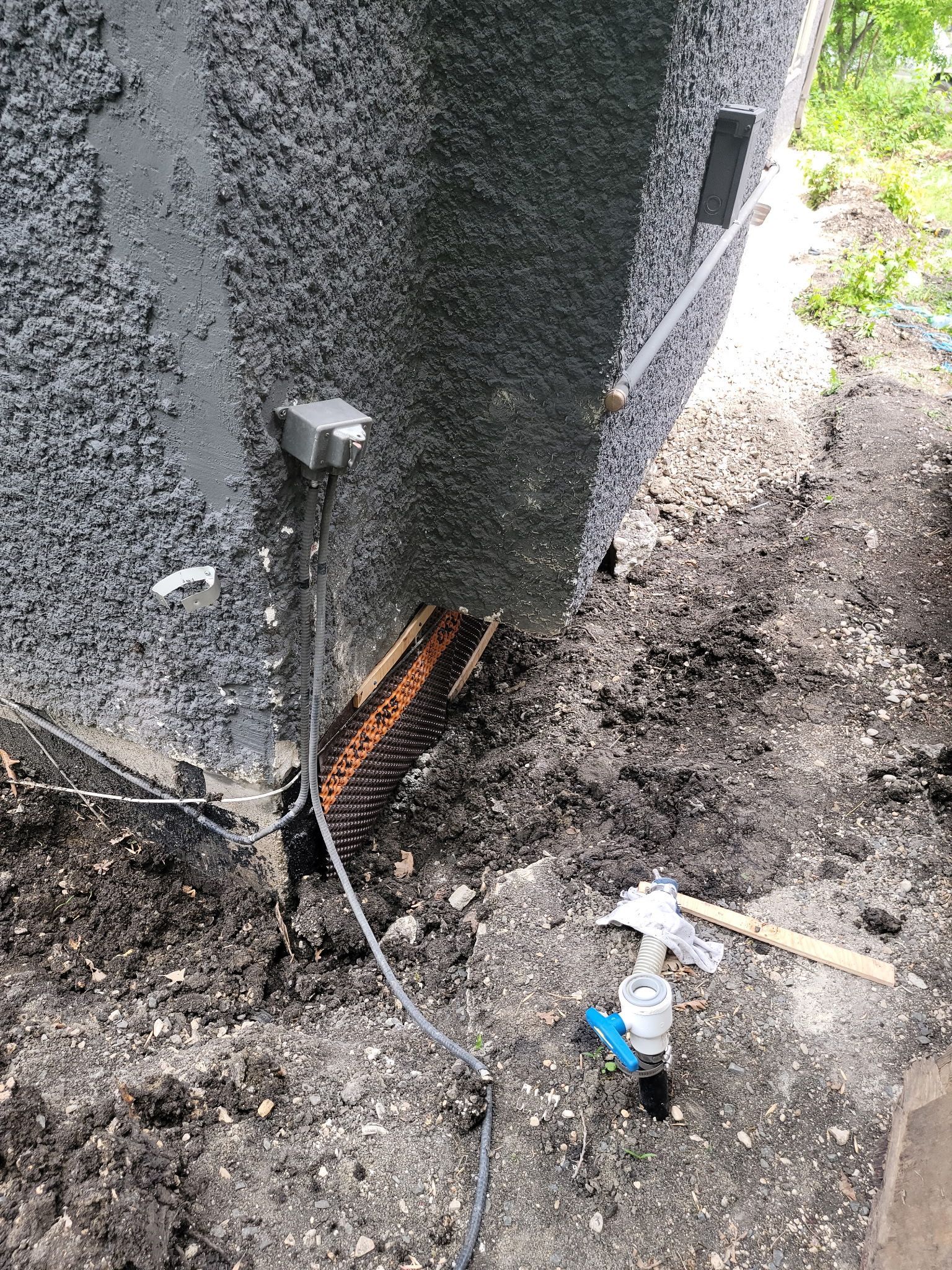 A dark, textured exterior house wall with an electrical junction box, a drainage strip at the base, and a spigot pipe.