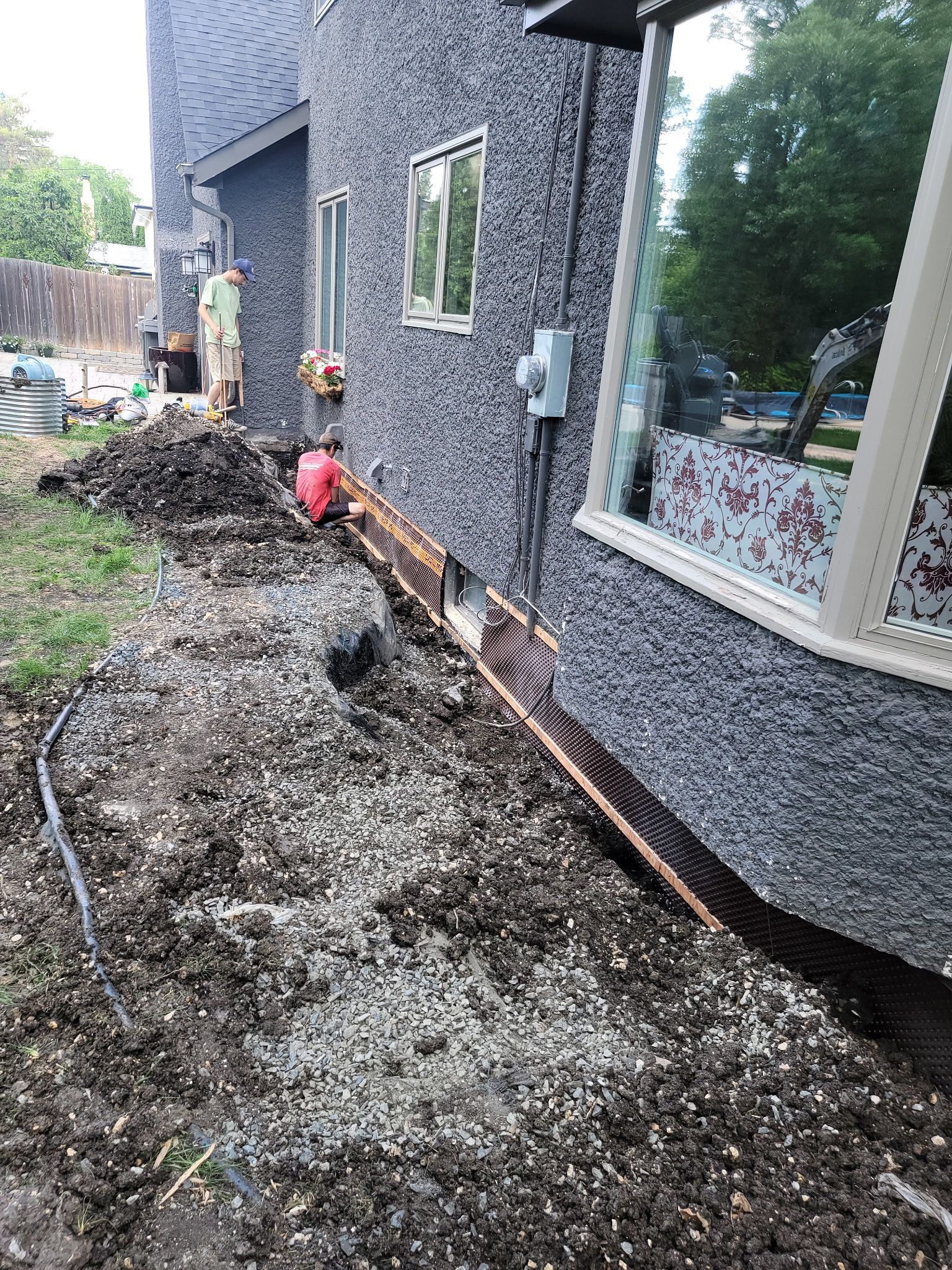 Two workers in a yard install a foundation drainage system along the exterior wall of a dark-textured house.