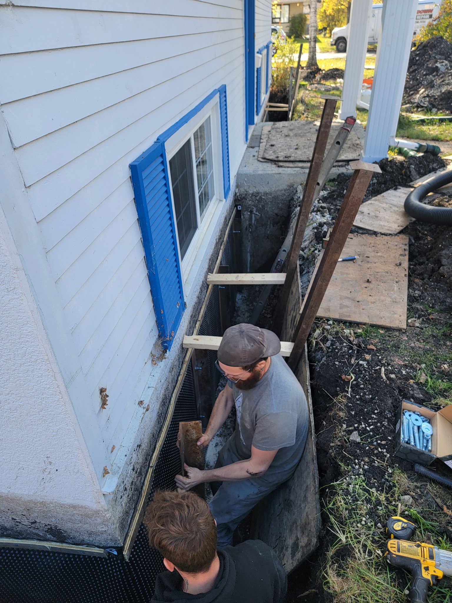 Two people work in a trench next to a white house, installing foundation waterproofing materials along the wall.