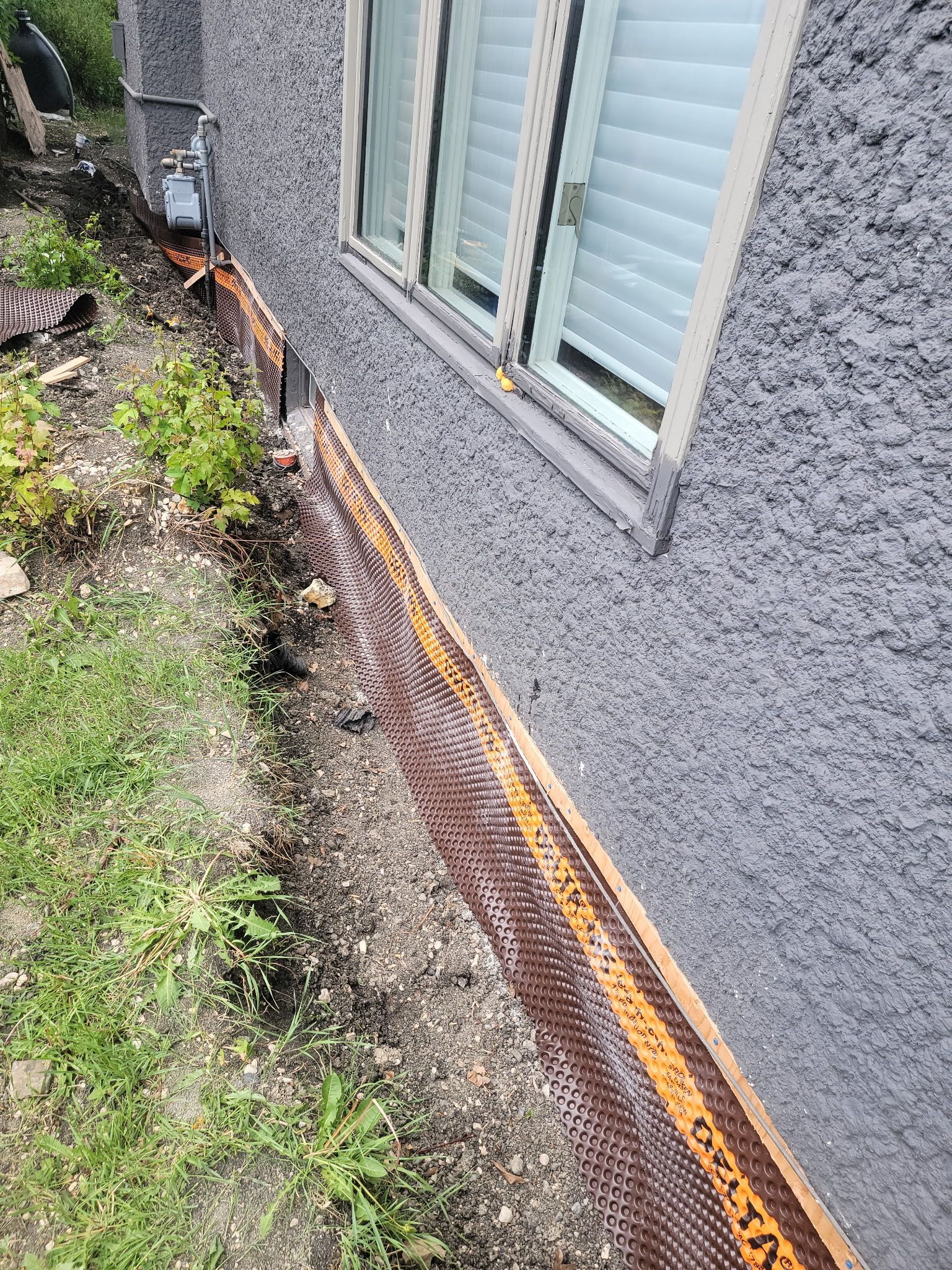 An orange protective drain board installed along the grey stucco foundation of a house exterior, above a gravel ground.