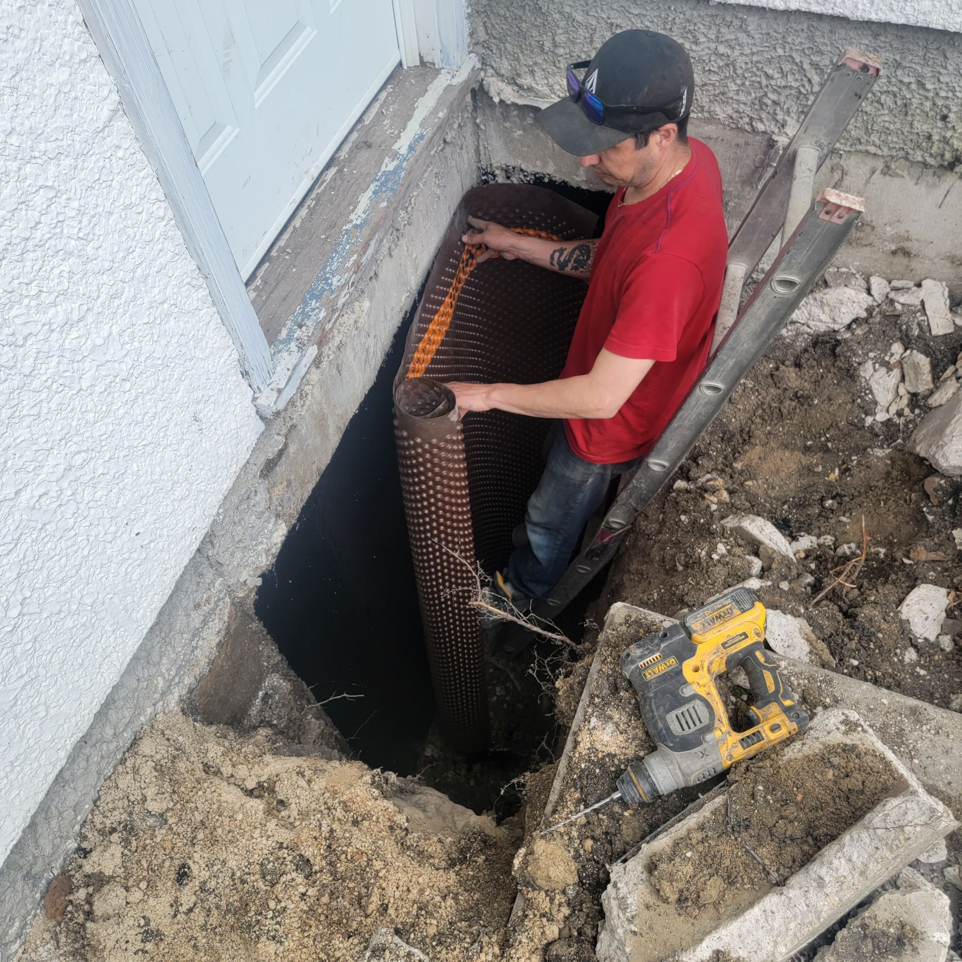 A worker installing a dimpled drainage membrane against a basement foundation wall in a deep excavation pit.