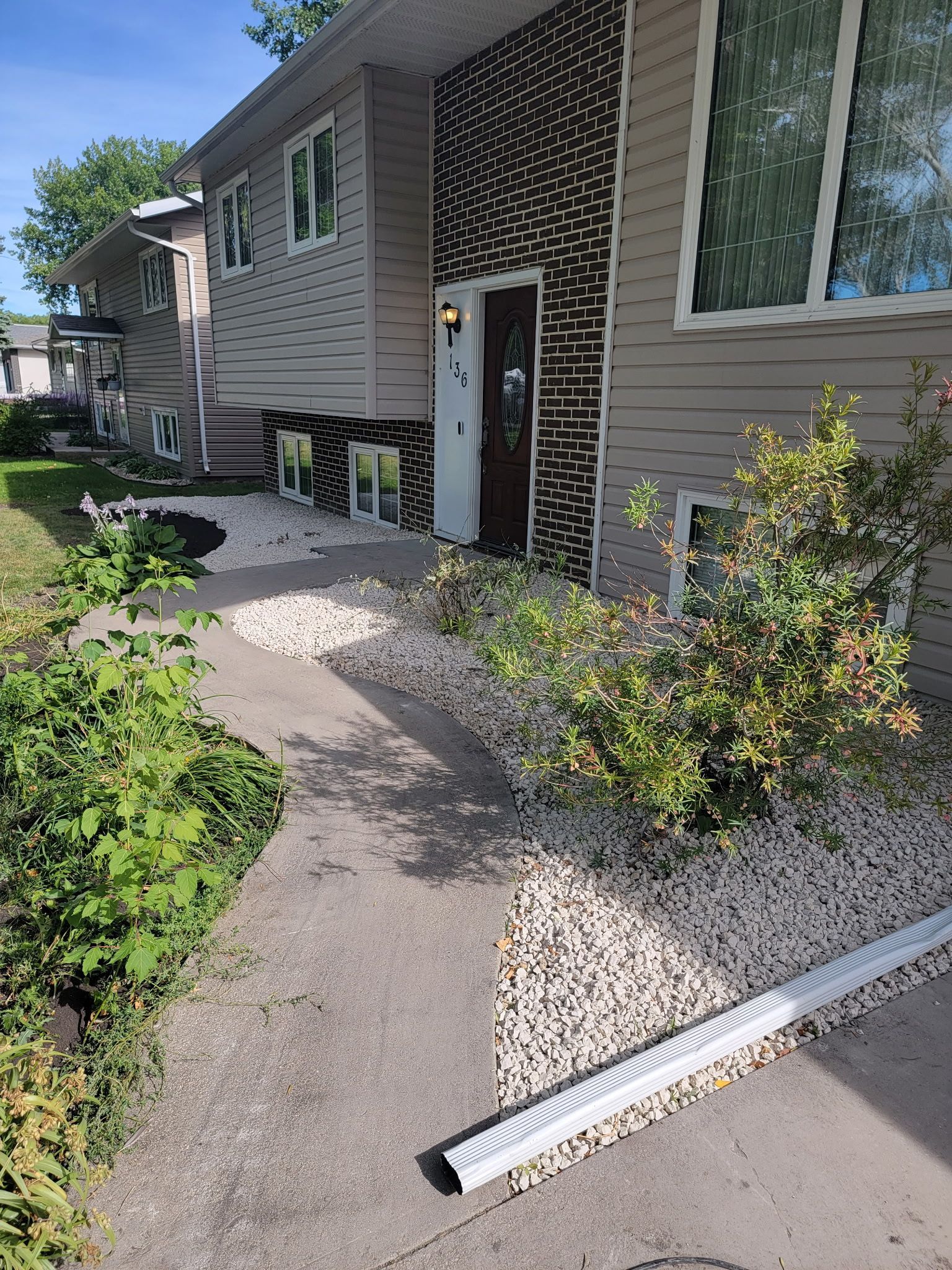 A split-level house exterior with beige siding, dark decorative brickwork, a front walkway, and a white downspout extension.