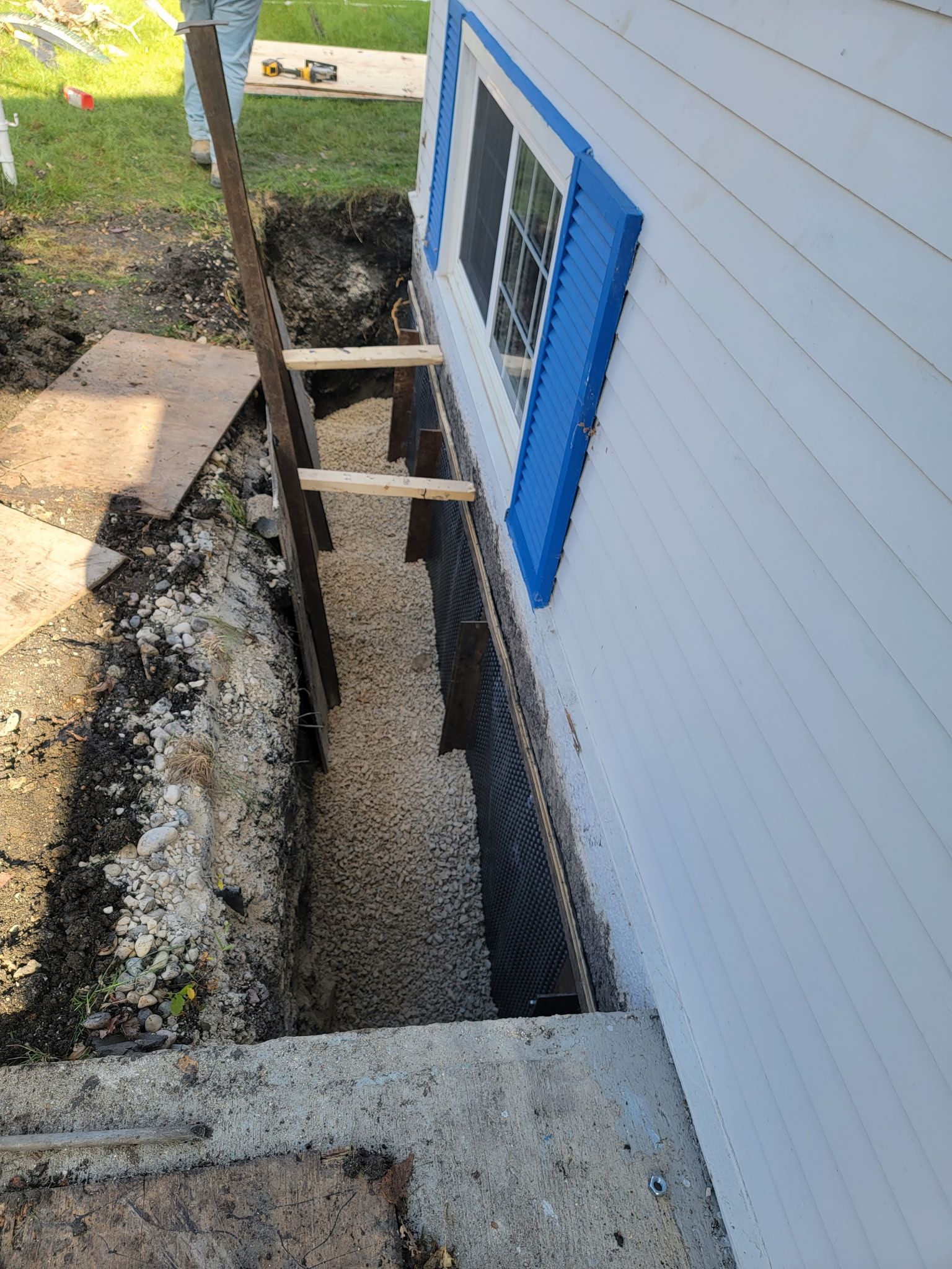 A foundation repair project showing a deep trench along a home exterior, featuring a drainage mat and gravel fill.