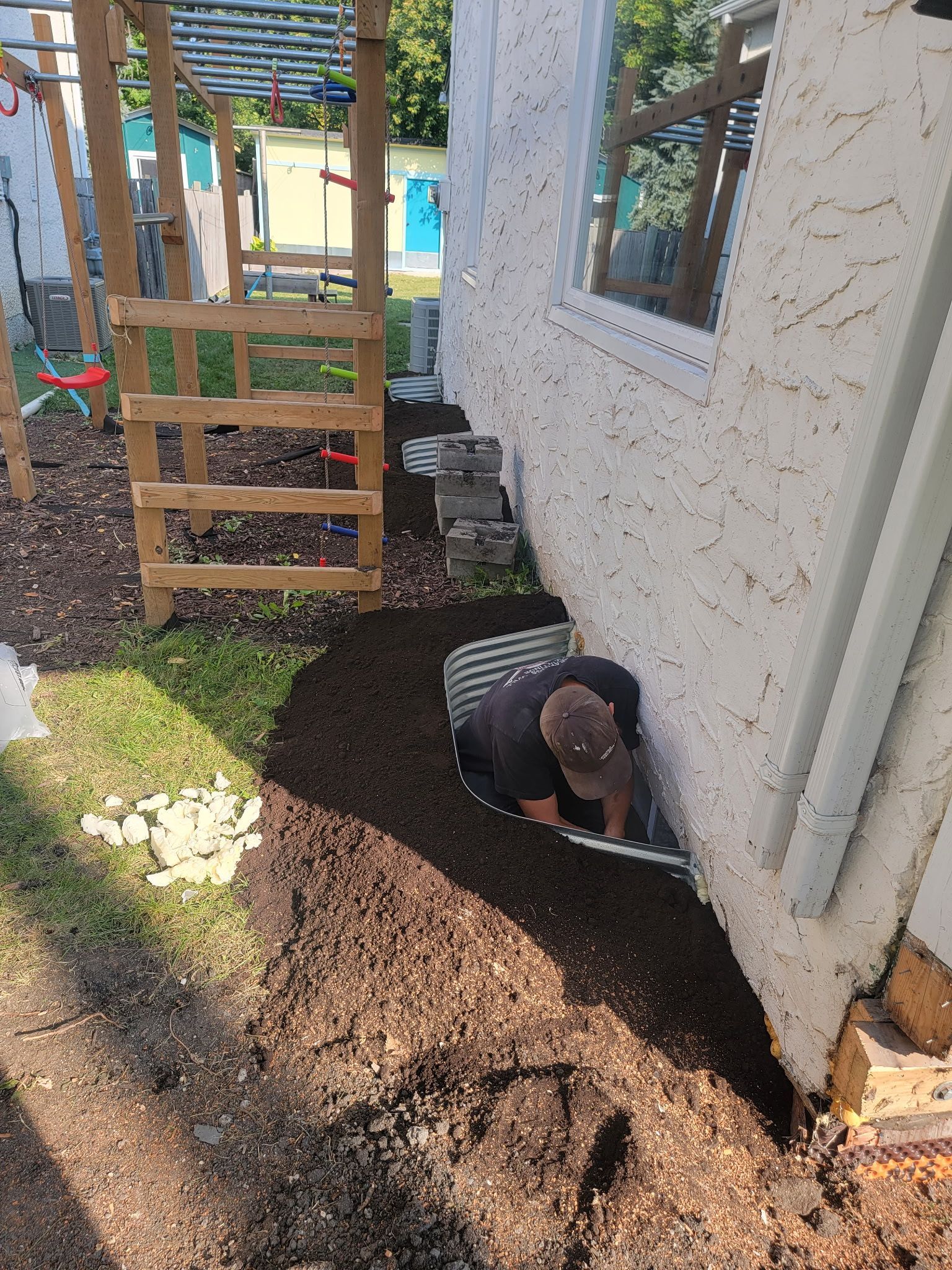 A person installs a corrugated metal window well against a stucco house wall surrounded by mulch and a wooden structure.