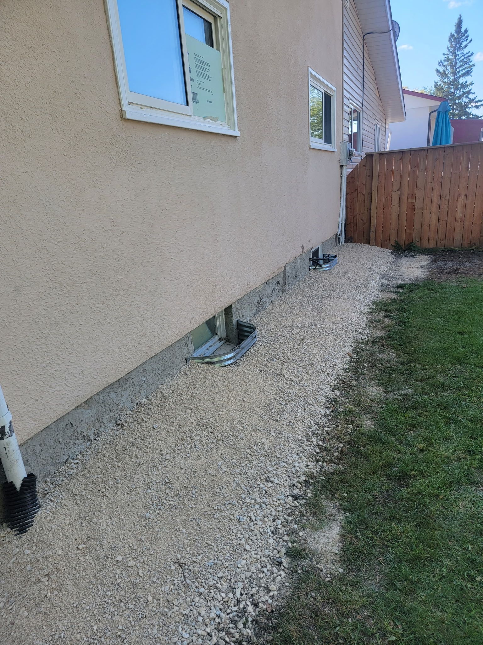 Tan house exterior with a gravel pathway along the foundation, a brown fence, and a green lawn under a sunny blue sky.