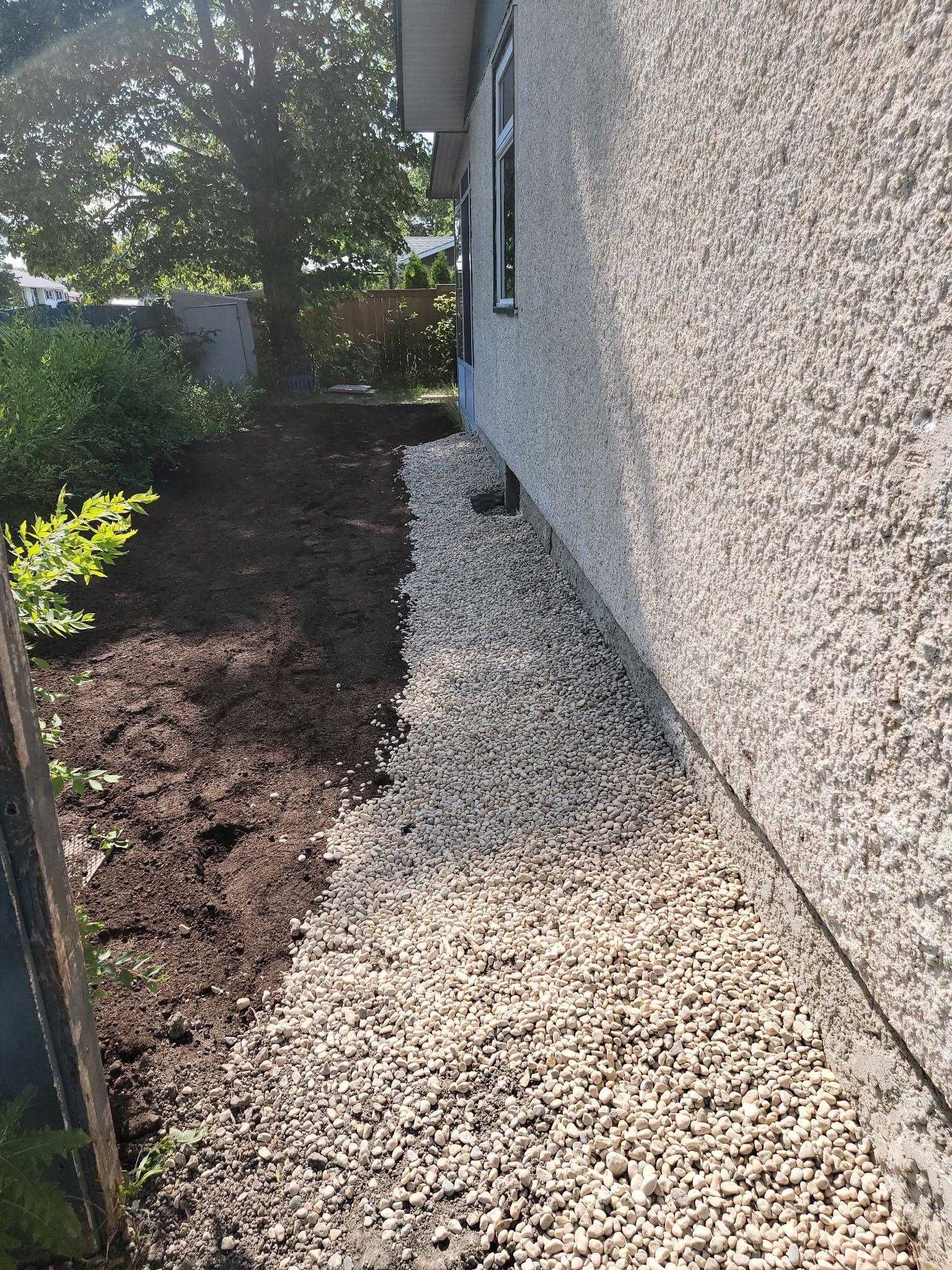A strip of light-colored gravel runs along the base of a pebble-dashed wall next to a patch of dark soil and trees.