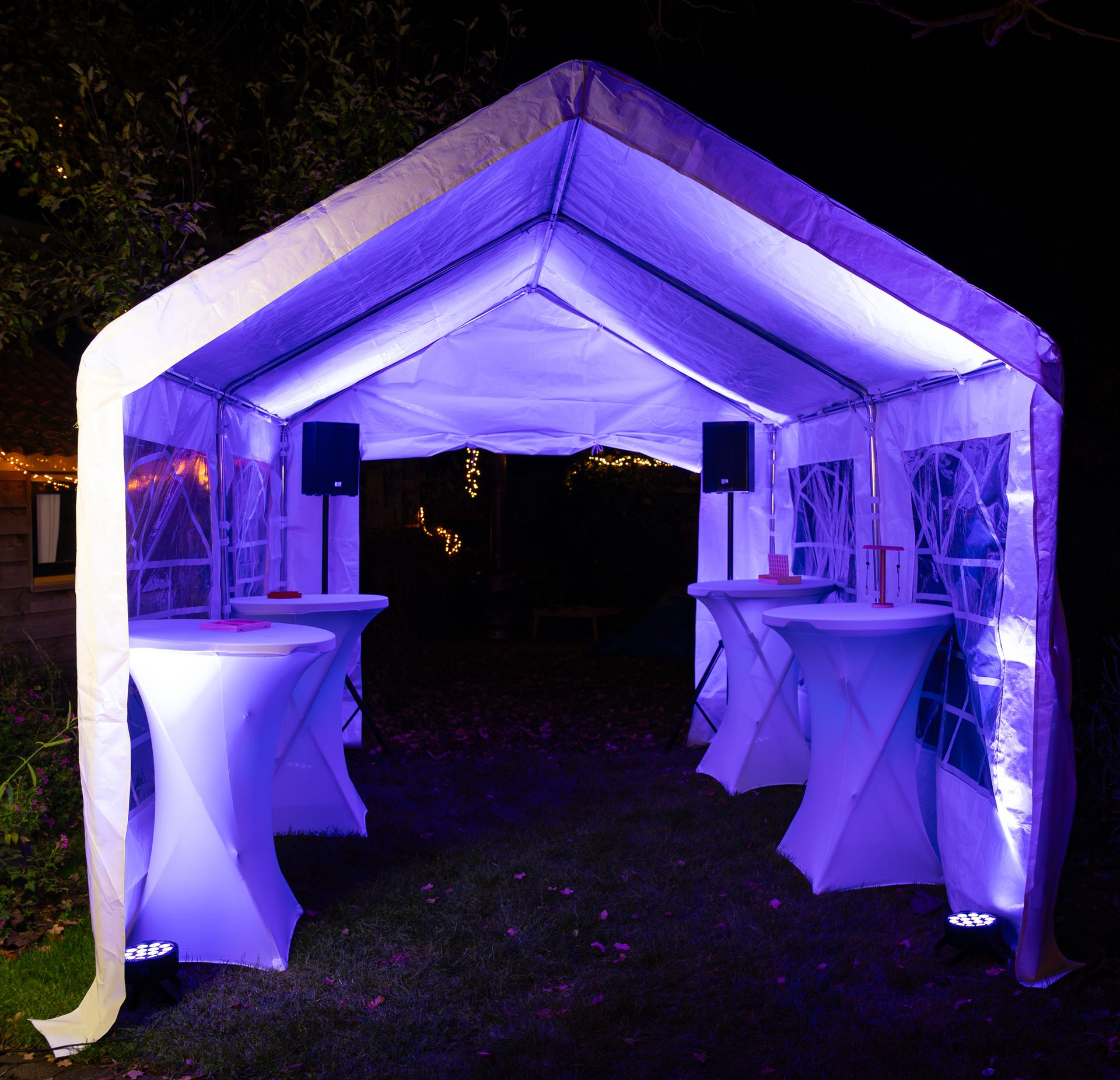 Een witte partytent, 's nachts verlicht met paarse uplighting, met hoge cocktailtafels en luidsprekers.