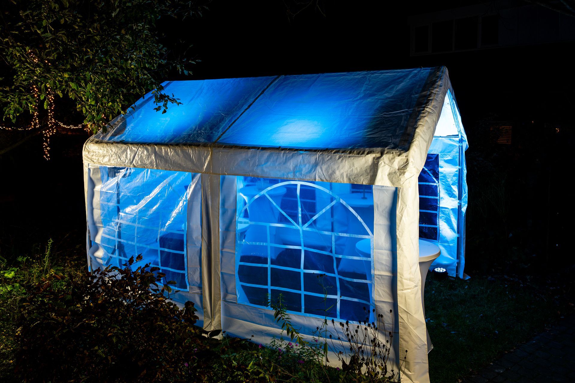 Een witte evenemententent voor buiten, 's nachts verlicht met een levendige blauwe gloed, staat op een grasveld te midden van donker gebladerte.