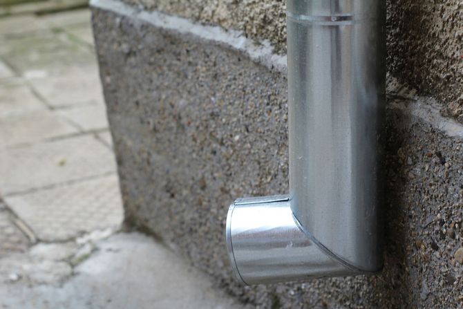 Chrome drainpipe elbow against a stone wall near a paved walkway