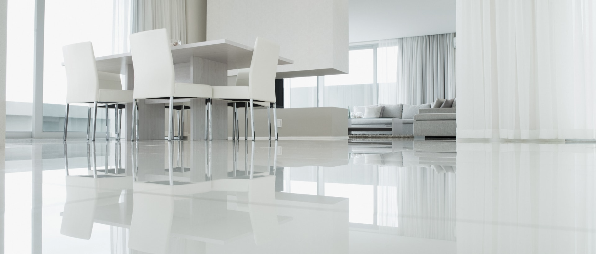 Modern white dining room with glass table, chairs, and glossy reflective floor near large windows