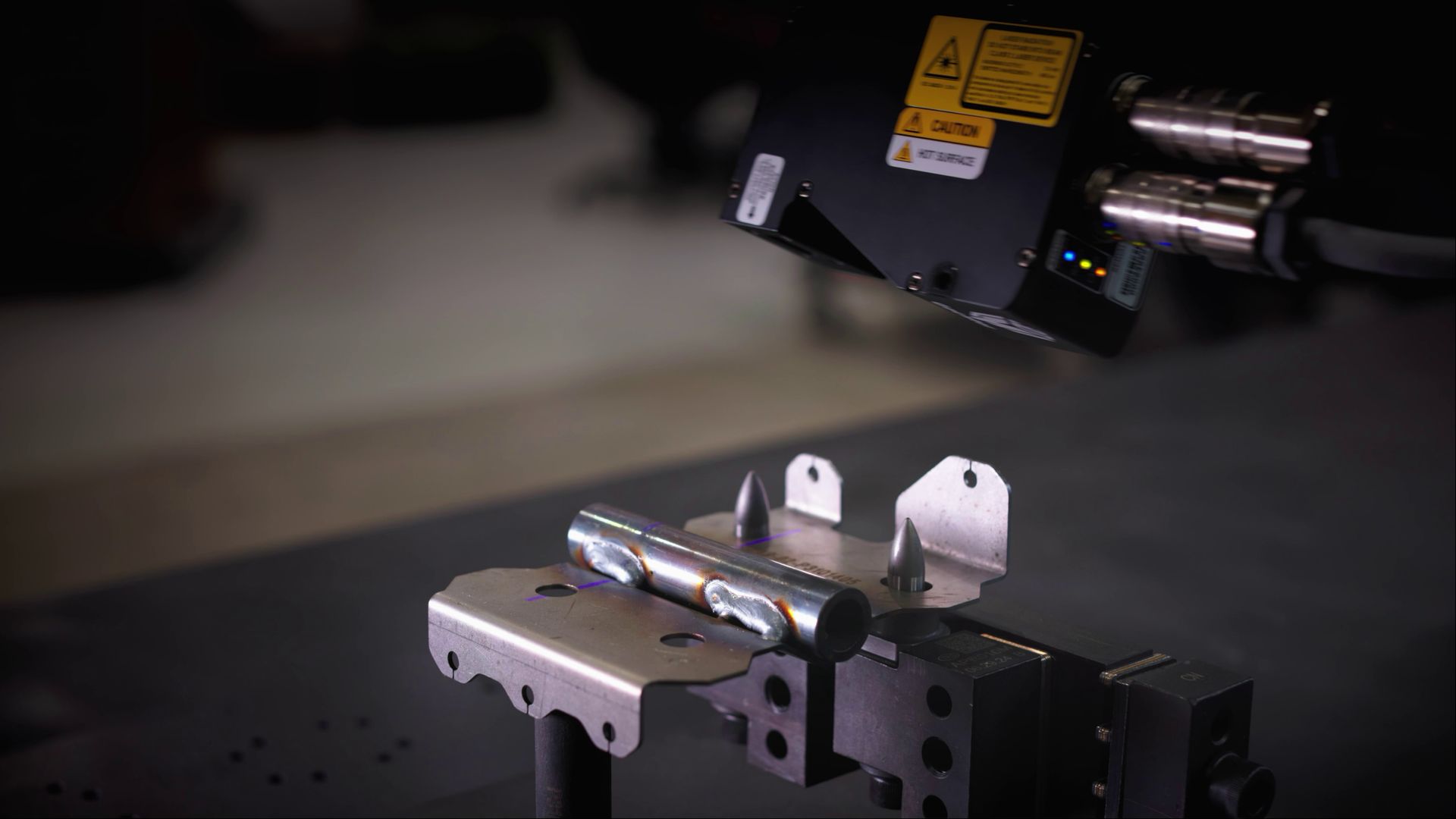 A robotic arm hovers over a small metallic fixture on a dark lab bench.