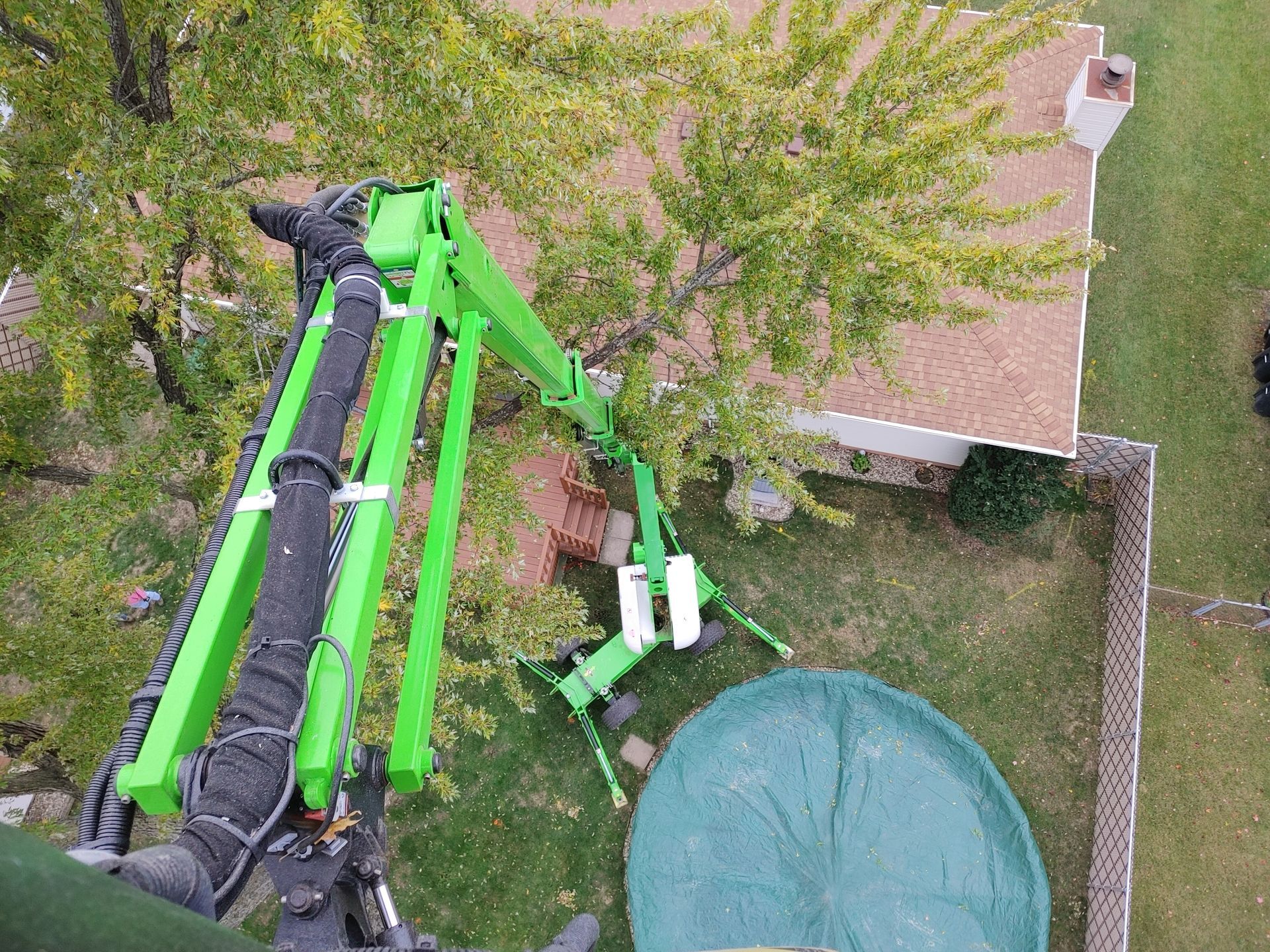An aerial view of a green crane lifting a tree in front of a house.