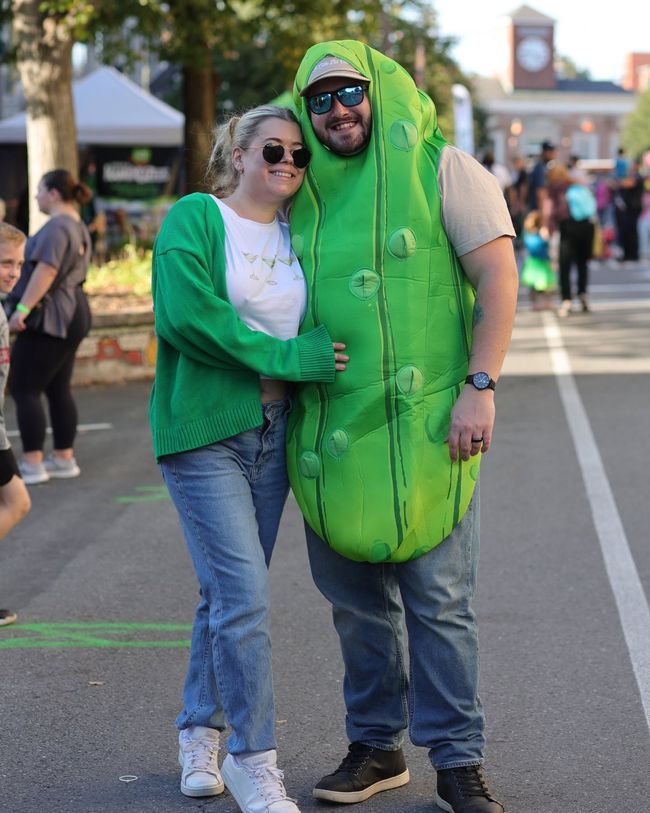 Two people in matching green pickle costumes posing and smiling outdoors on a street.