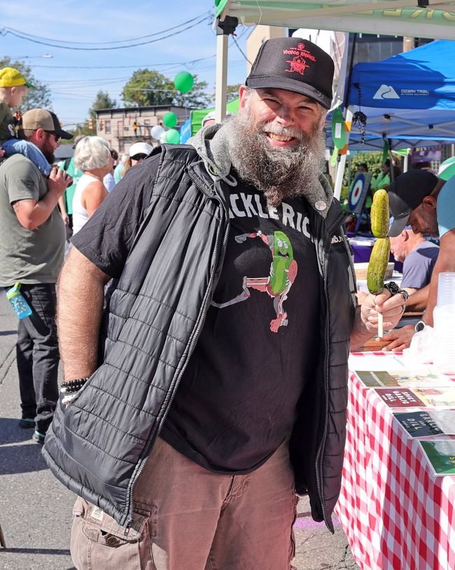 Man with beard in black cap and vest standing at an outdoor fair beside a red-checkered table