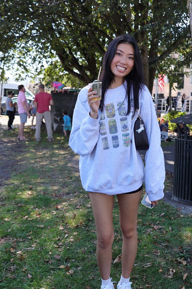 Smiling person in a white hoodie and shorts standing outdoors in a park near trees and grass.