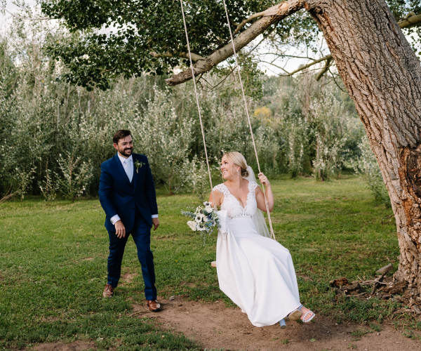 Bride on a swing under a tree, smiling at groom in navy suit in a grassy garden.