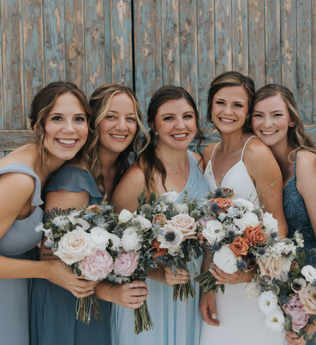 Five women holding pastel bridal bouquets in front of a weathered wooden wall, smiling at the camera