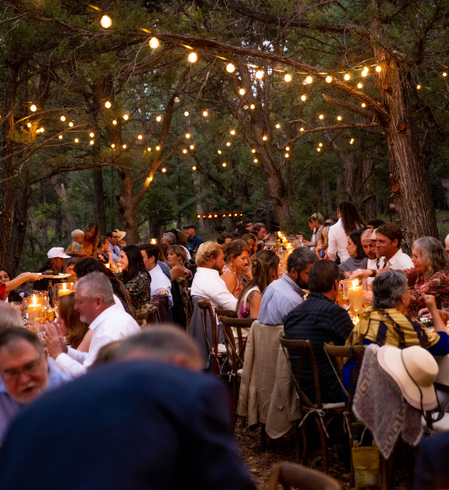 Outdoor dinner party under string lights in a wooded setting, with guests seated at long tables.