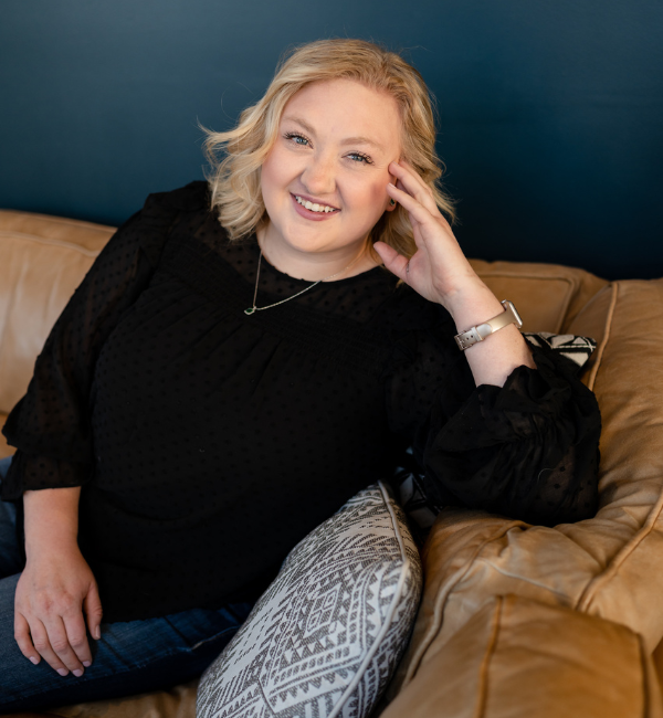 Smiling person reclining on a tan couch against a dark blue wall, wearing a black top.